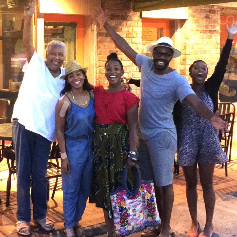 Five smiling people, two men and three women, with arms raised, standing outside a restaurant at dusk.