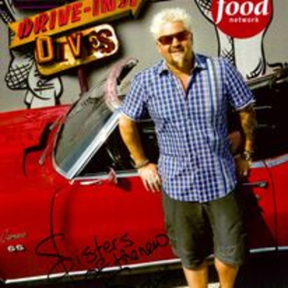 Guy Fieri in sunglasses and a plaid shirt, leaning on a red classic car with a "Drive-Ins, Diners, and Dives" sign.