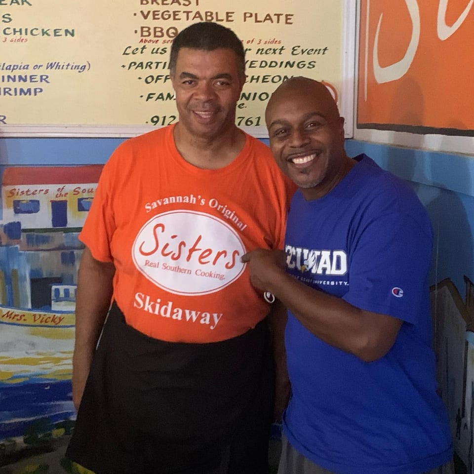 Two smiling men, one in an orange "Sisters Real Southern Cooking" shirt, stand in a restaurant.