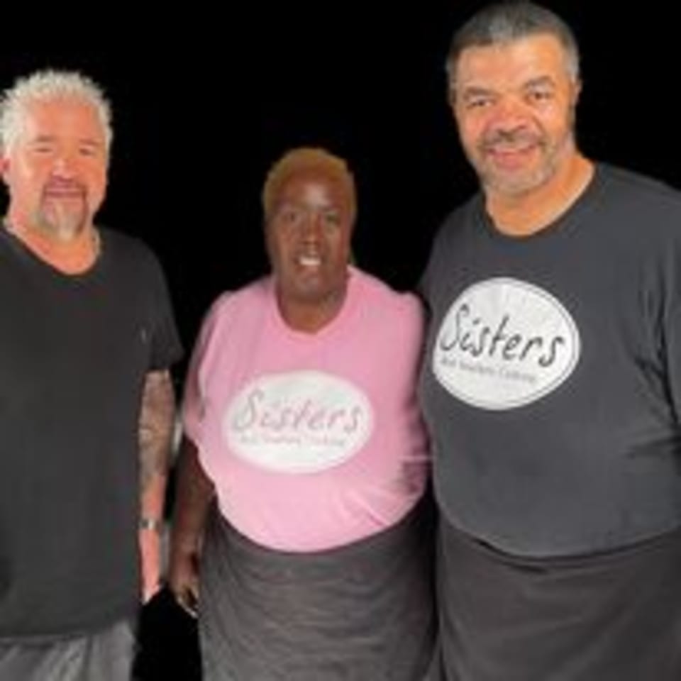 Guy Fieri poses with two Sisters Soul Food Catering staff members in branded shirts.